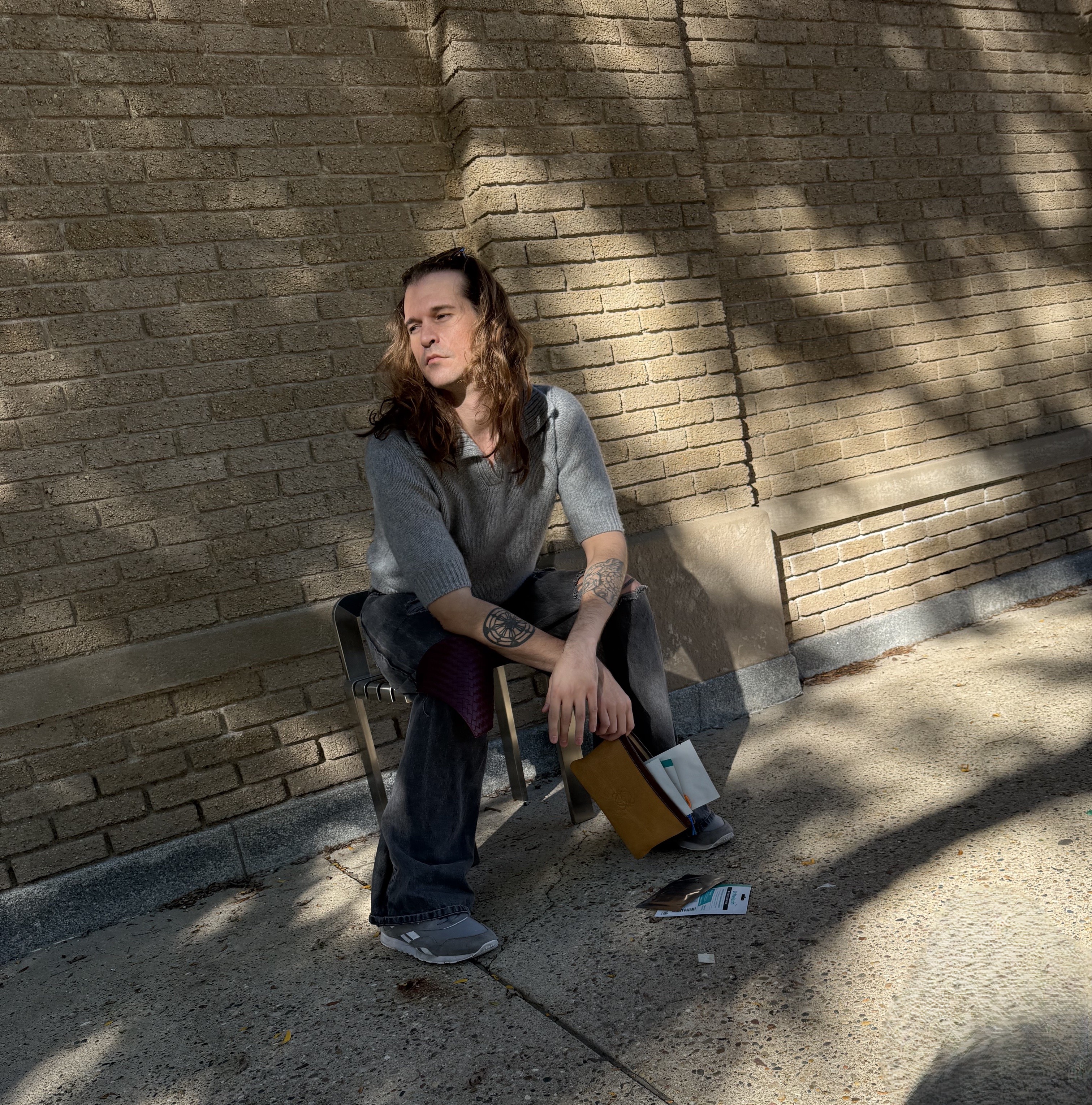 Seated on a chair against the sunlit brickwall, grey tee, baggy jeans