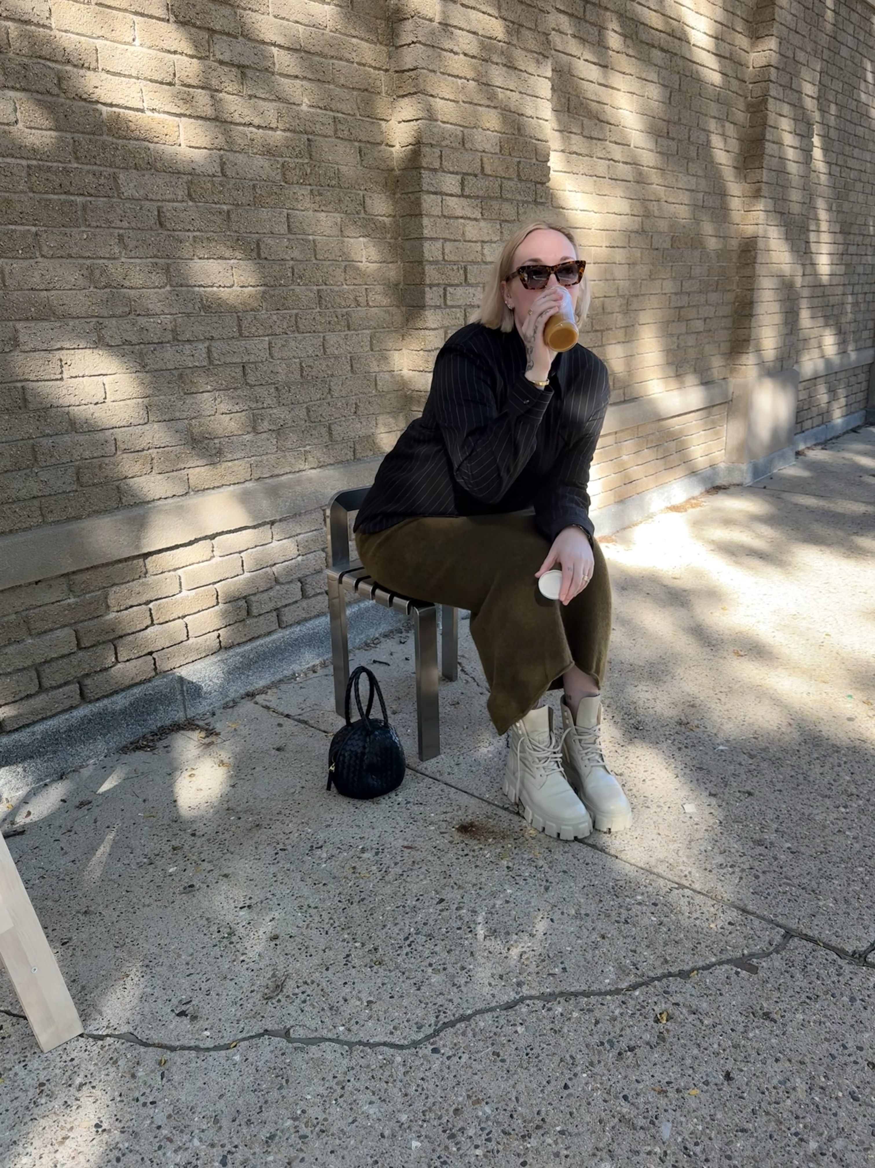 Sunglasses, black top, olive trousers, seated, small bag at her feet